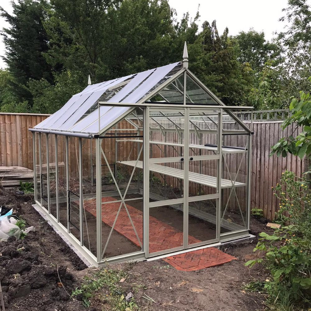 A Rhino Greenhouse with white framework and transparent panels stands on a red brick path, surrounded by soil and greenery, enclosed by a wooden fence in a garden setting.