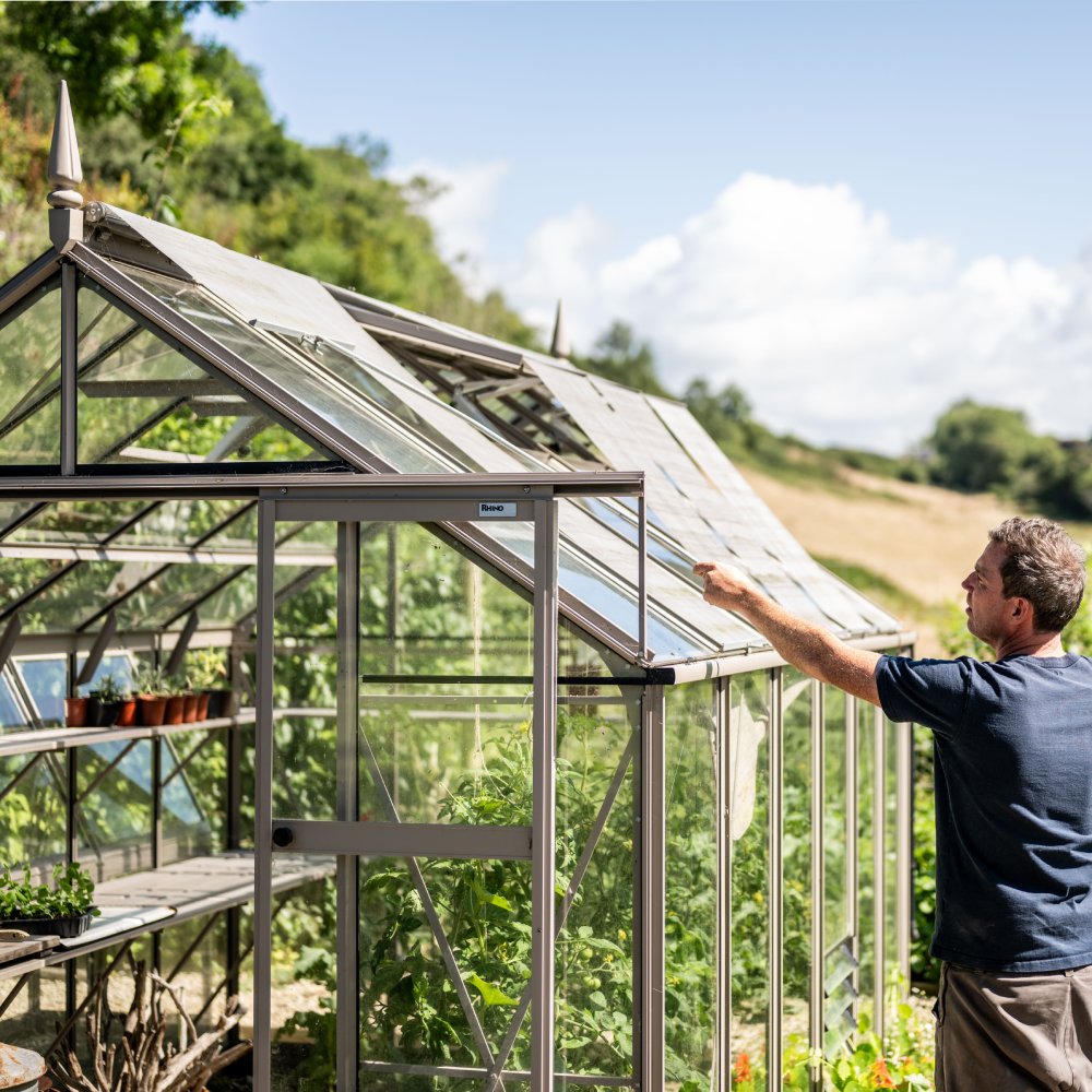 A man adjusts a vent on a large, glass-paneled Rhino Greenhouse labeled Rhino in a lush, rural landscape. Potted plants sit on shelves inside under a sunny, blue sky.