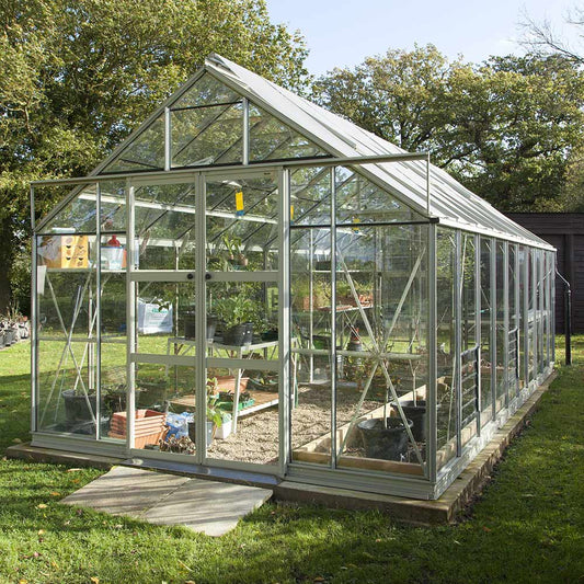 A glass Rhino Greenhouse stands in a grassy garden, housing various potted plants and gardening tools. Sunlight filters through the transparent panels, surrounded by trees and natural greenery.