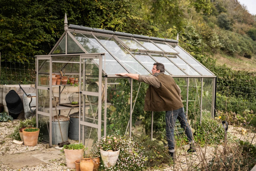 A man opens a glass Rhino Greenhouse door, revealing potted plants and gardening tools inside. The Rhino Greenhouse is set in a garden with lush greenery around it.