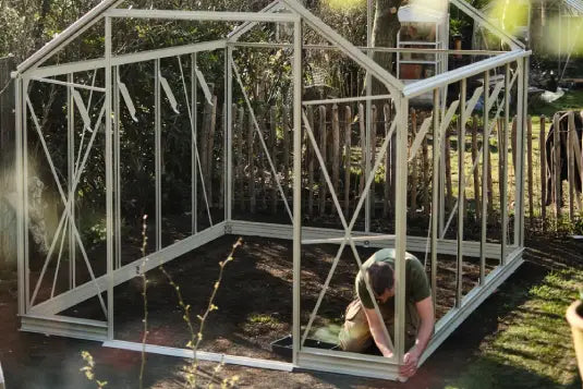 A person assembles a metal Rhino Greenhouse frame in a garden, surrounded by plants and a wooden fence under sunlight.