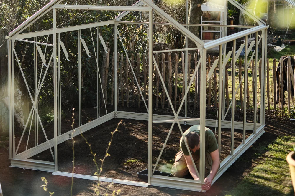 A man assembles the frame of a Rhino Greenhouse in a garden. Sunlight filters through trees, and a wooden fence surrounds the area, creating a serene, natural setting.