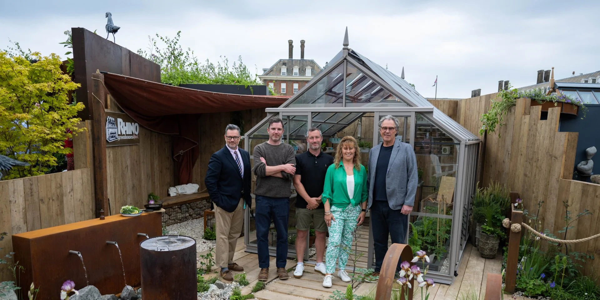 A group of five people stands in front of a metal Rhino Greenhouse, surrounded by wooden fencing and plants. A sign reads, Rhino Rhino Greenhouses Direct. A sculpture and water feature are visible nearby.