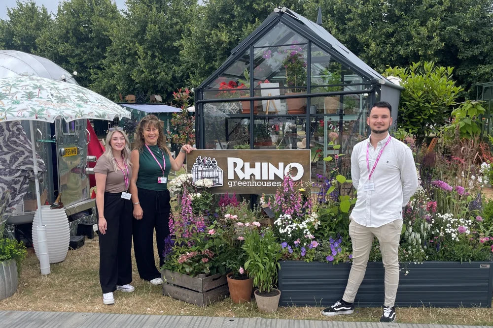 A small glass Rhino Greenhouse with plants inside and around, surrounded by three people on grass. A RHINO Rhino Greenhouses sign is displayed in front of colorful flowers and greenery.