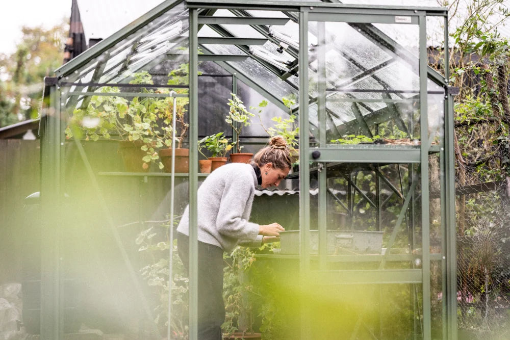 A person tends to plants in a glass Rhino Greenhouse, surrounded by pots and greenery. The setting is outdoors with trees and bushes visible in the background.