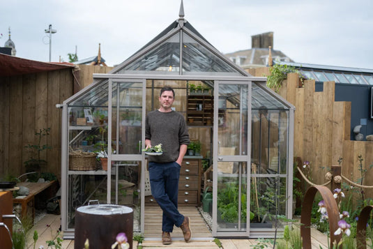 A person stands holding plants in front of a glass Rhino Greenhouse filled with gardening tools and plants, surrounded by wooden fencing and outdoor decor.