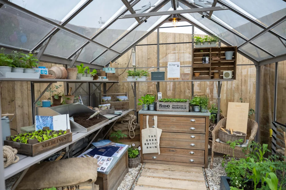 Inside a wooden Rhino Greenhouse, plants thrive on shelves and a wooden table. Gardening tools and seed packets are neatly organized. A sign reads, RHINO Rhino Greenhouses Handmade in NORFOLK.
