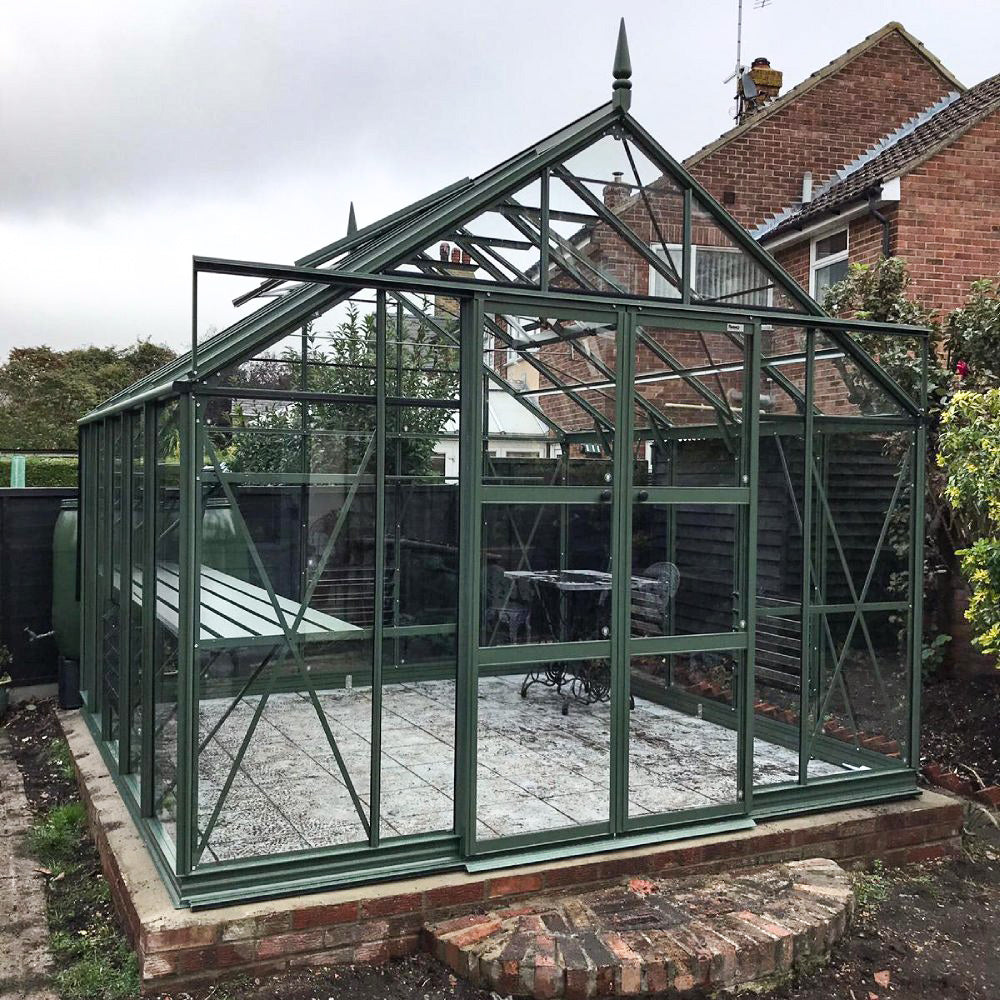 Rhino Greenhouse with dark green metal framing and glass panels is unoccupied, standing on a brick foundation. It is nestled in a garden with surrounding trees and adjacent brick houses.