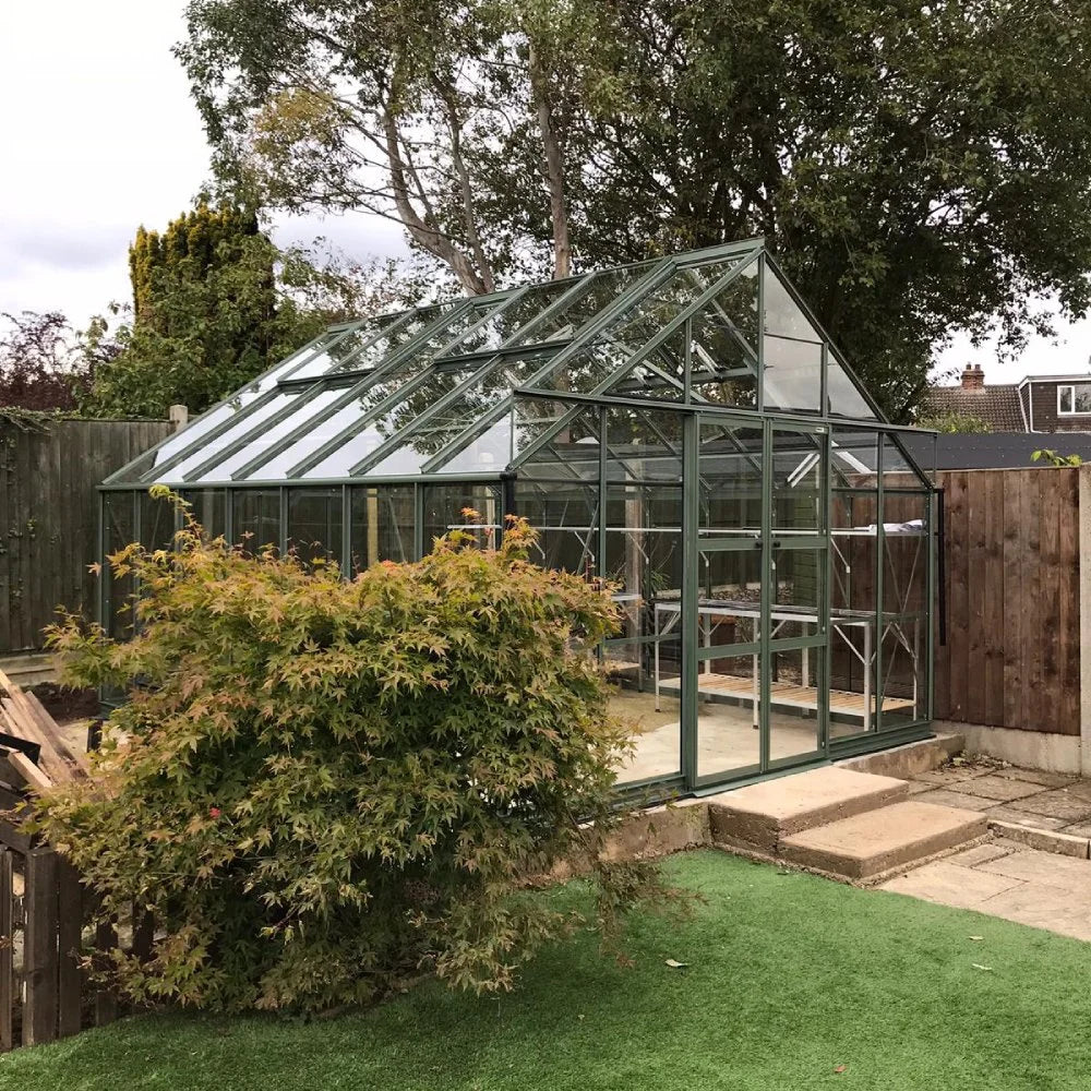 Rhino Greenhouse with a glass-paneled structure stands on a stone patio. It is surrounded by a wooden fence and lush trees and bushes, within a garden setting.