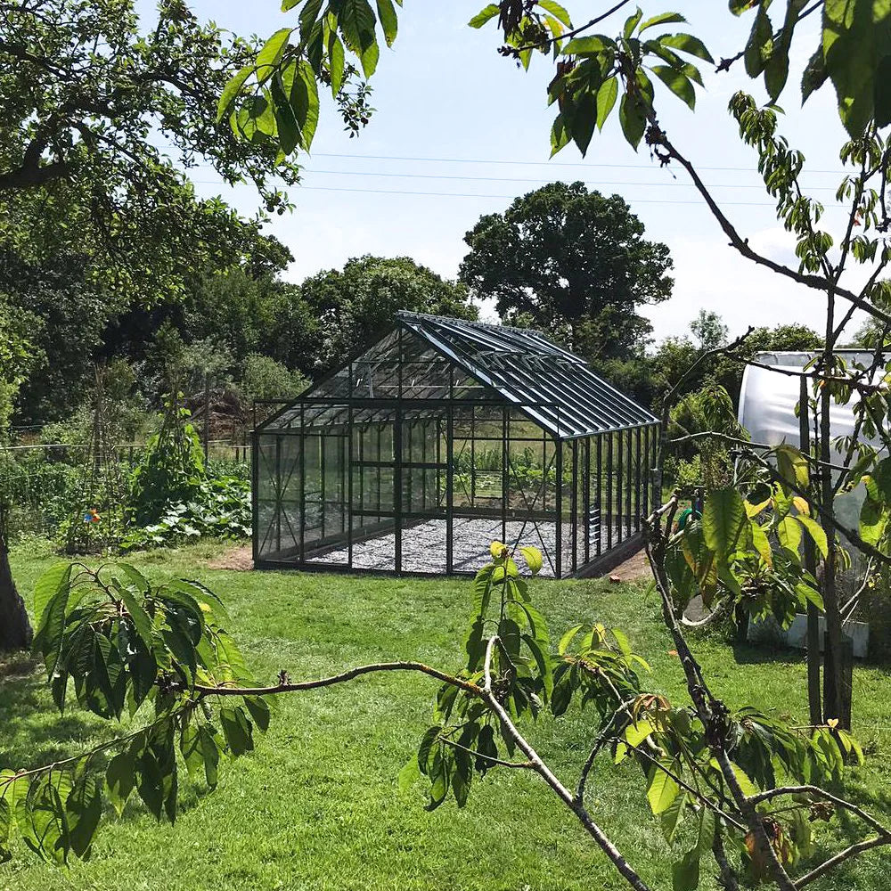 A glass Rhino Greenhouse stands on a grassy lawn, surrounded by trees and plants. Sunlight reflects off the structure, highlighting its geometric frame in a lush garden setting.