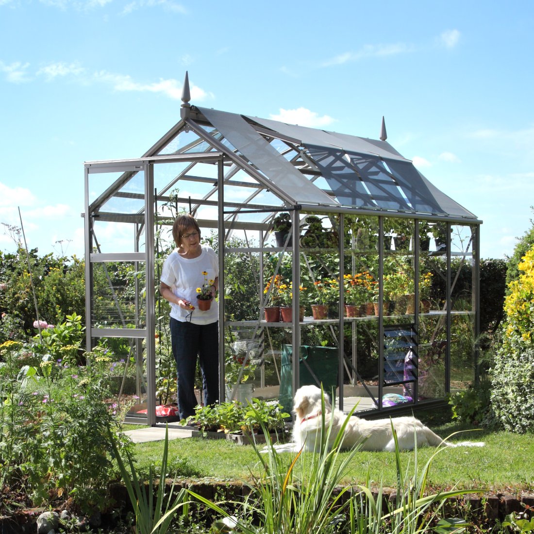 A glass Rhino Greenhouse houses potted plants, with a woman tending to a flowerpot. A dog lounges on nearby grass, surrounded by lush garden foliage under a bright blue sky.