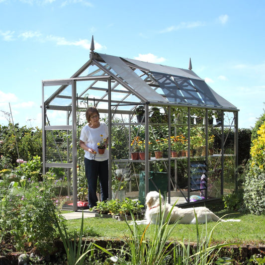 A glass Rhino Greenhouse houses potted plants, with a woman tending to a flowerpot. A dog lounges on nearby grass, surrounded by lush garden foliage under a bright blue sky.