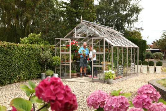 A Rhino Greenhouse with two people inside is surrounded by a lush garden. They appear to be tending to plants. Vibrant flowers and neatly trimmed hedges decorate the gravel pathway.