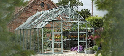A glass Rhino Greenhouse stands in a garden, displaying colorful plants inside on shelves. It is set against a backdrop of a red-brick building and surrounded by greenery.