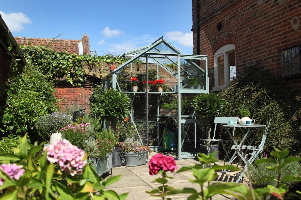 A glass Rhino Greenhouse stands surrounded by blooming flowers and potted plants. A small seating area with a table and chairs is placed nearby, all in a sunlit garden courtyard.