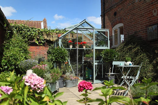 A glass Rhino Greenhouse stands surrounded by blooming flowers and potted plants. A small seating area with a table and chairs is placed nearby, all in a sunlit garden courtyard.