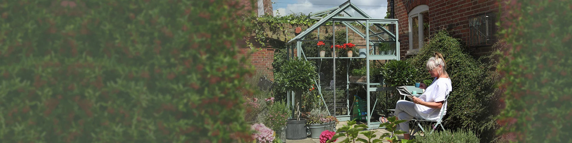 A Rhino Greenhouse stands amidst a lush garden, surrounded by brick walls. A person sits nearby, reading, surrounded by potted plants and flowers, enjoying a serene outdoor setting.