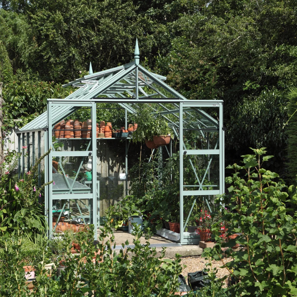 A glass Rhino Greenhouse stands surrounded by lush greenery, containing potted plants and gardening tools, with terracotta pots lined up inside. It's set in a garden with trees in the background.