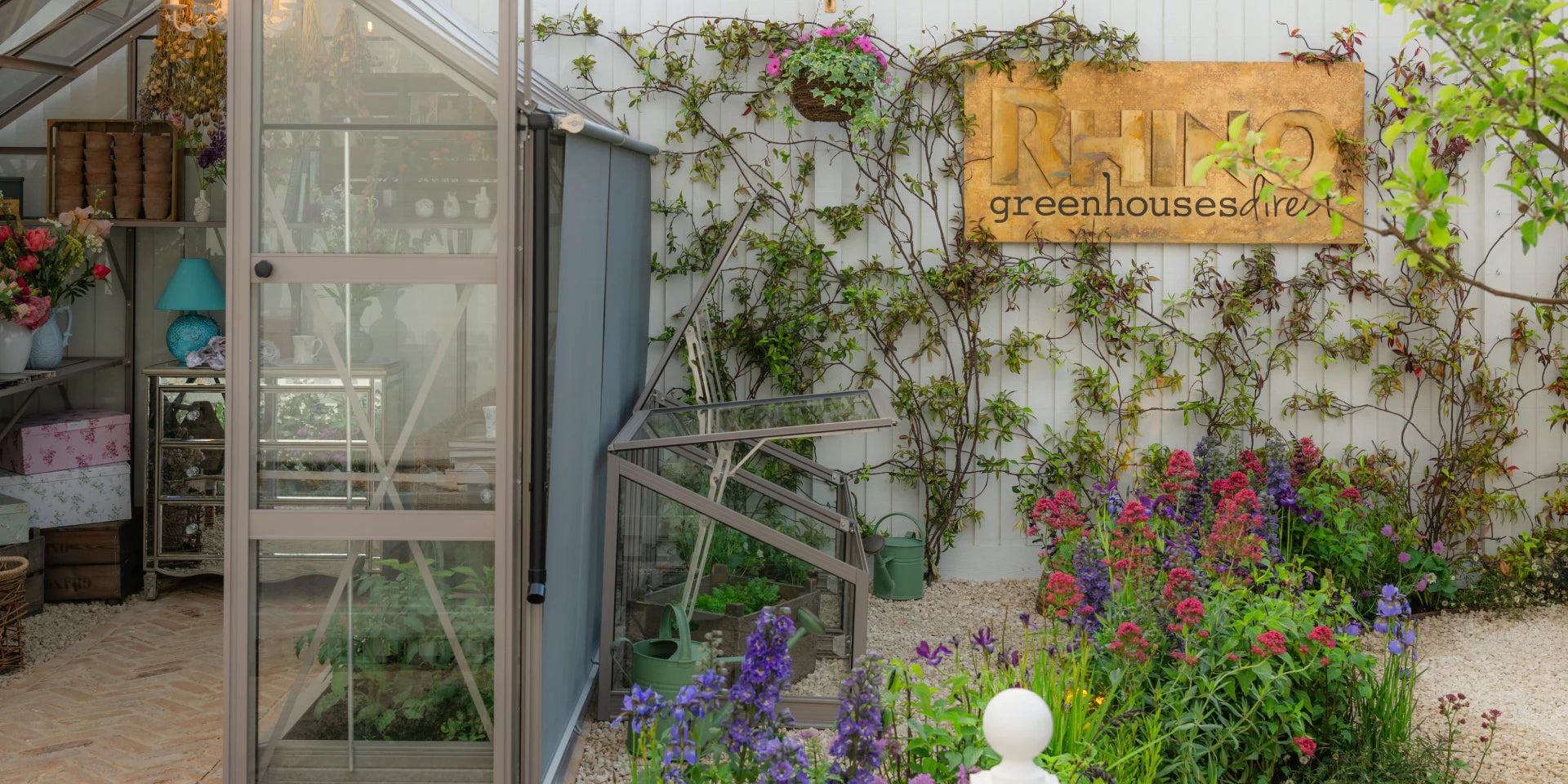 Glass Rhino Greenhouse filled with plants and gardening tools, situated next to a wall covered in climbing vines and colorful flowers. A sign reads RHINO Rhino Greenhouses direct.
