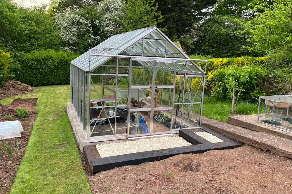 A glass Rhino Greenhouse stands on a grassy lawn, filled with gardening tools and plants. Surrounding it are lush green hedges and trees, creating a serene, natural garden setting.