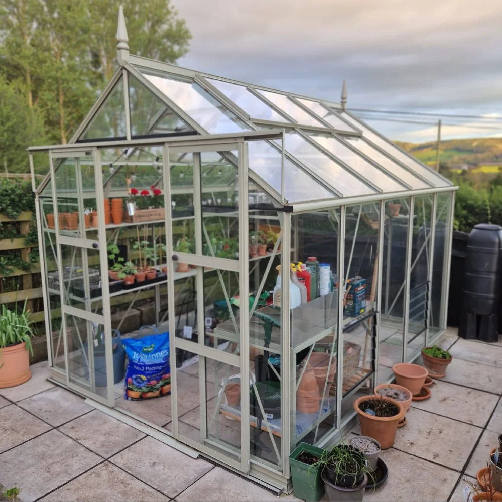 A glass Rhino Greenhouse, filled with potted plants and gardening supplies, stands on a tiled patio surrounded by greenery under a cloudy sky. Nearby, terracotta pots and garden tools are visible.