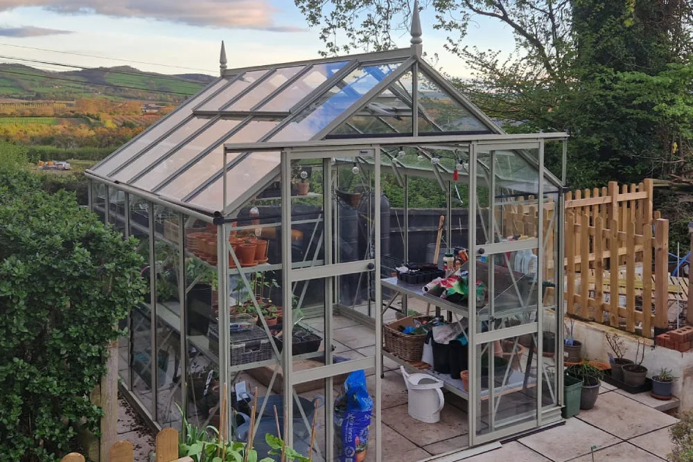 A glass Rhino Greenhouse filled with potted plants and gardening tools stands on a paved area. Surrounded by lush greenery and wooden fencing, it overlooks rolling hills under a partly cloudy sky.