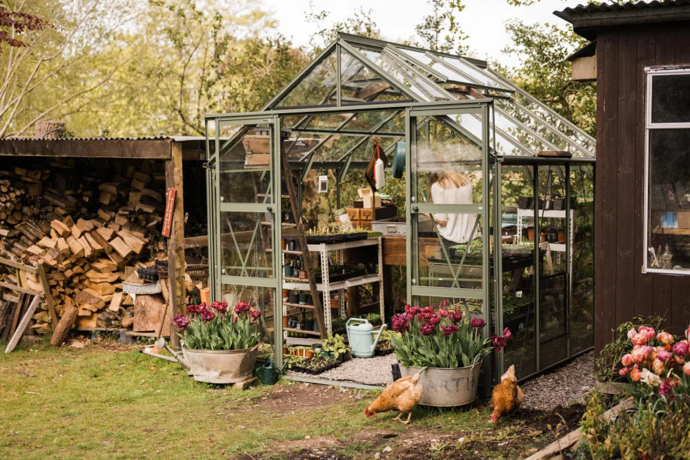 A glass Rhino Greenhouse with a person working inside is surrounded by wooden logs and colorful flowers. Chickens roam the grassy area outside.
