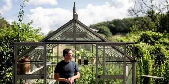 A person stands in front of a large glass Rhino Greenhouse with a pointed roof, surrounded by lush greenery and a partly cloudy sky.