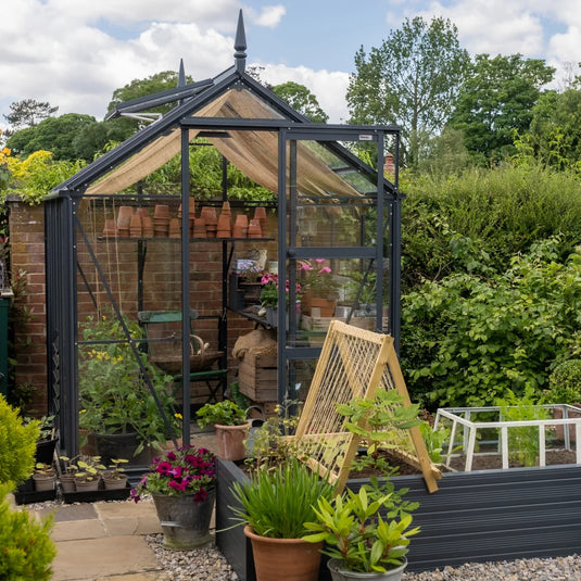 A glass Rhino Greenhouse contains various potted plants and gardening tools, with additional plants and a small trellis outside. It is set against a lush garden backdrop with trees and hedges.