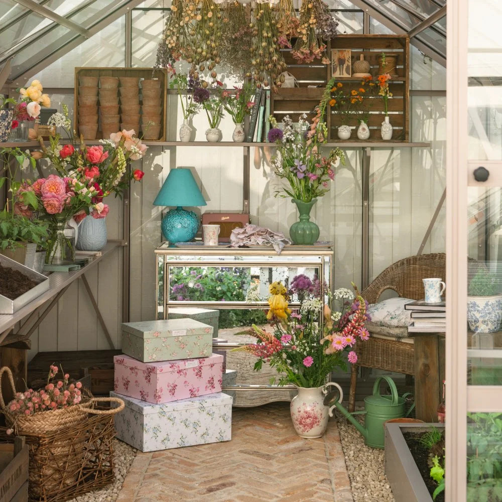 A Rhino Greenhouse interior features a variety of colorful flowers in vases and baskets on shelves and tables, with gardening tools. Soft light filters through the glass structure, creating a serene atmosphere.