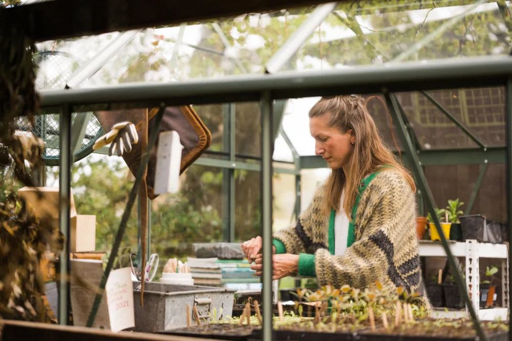 A person tends to seedlings inside a Rhino Greenhouse, wearing a patterned sweater. Surrounding them are plants, gardening tools, and gloves. The Rhino Greenhouse exterior reveals trees and foliage outside.