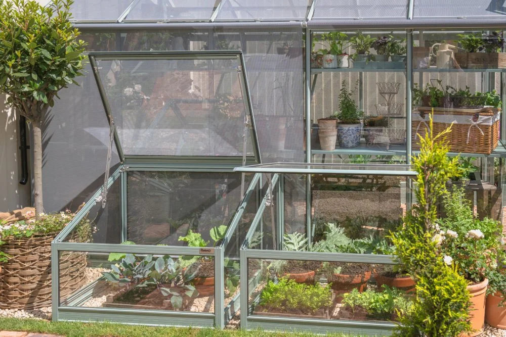Glass Rhino Greenhouse featuring open roof panels, housing various potted plants and vegetables, surrounded by lush garden greenery. Text on Rhino Greenhouse: Rhino.