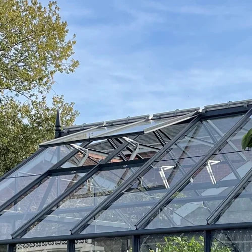A glass Rhino Greenhouse with open top vents lets in sunlight, surrounded by trees and a clear blue sky.