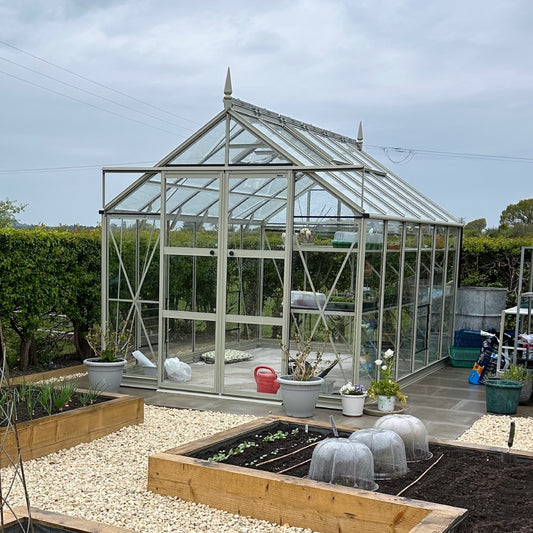 A glass Rhino Greenhouse stands in a garden, surrounded by raised wooden beds and gravel paths. Potted plants and gardening tools are visible nearby, creating a well-maintained planting area.