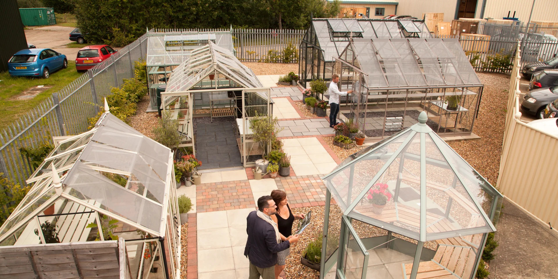 Greenhouses are displayed in an outdoor showroom. Two people examine a brochure, while another person inspects a Rhino Greenhouse. The area is neatly arranged with potted plants and cars parked nearby.