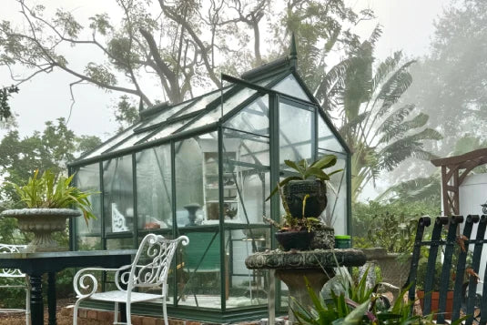 A glass Rhino Greenhouse stands amidst garden furniture and potted plants, surrounded by lush greenery and trees, in a misty outdoor setting.