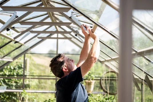 A person adjusts a window in a Rhino Greenhouse, surrounded by lush greenery and filtered sunlight, ensuring optimal conditions for plant growth.