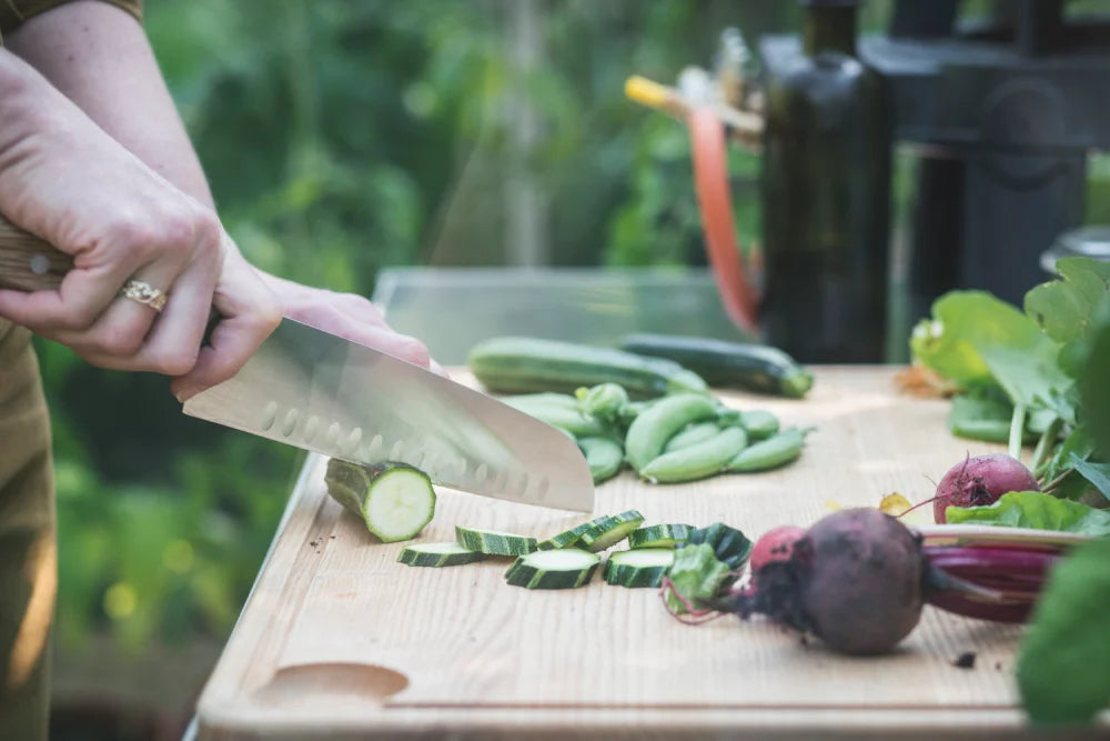 Hand slices cucumber with large knife on wooden cutting board, surrounded by fresh vegetables like snap peas and beets, in a garden setting with green foliage.
