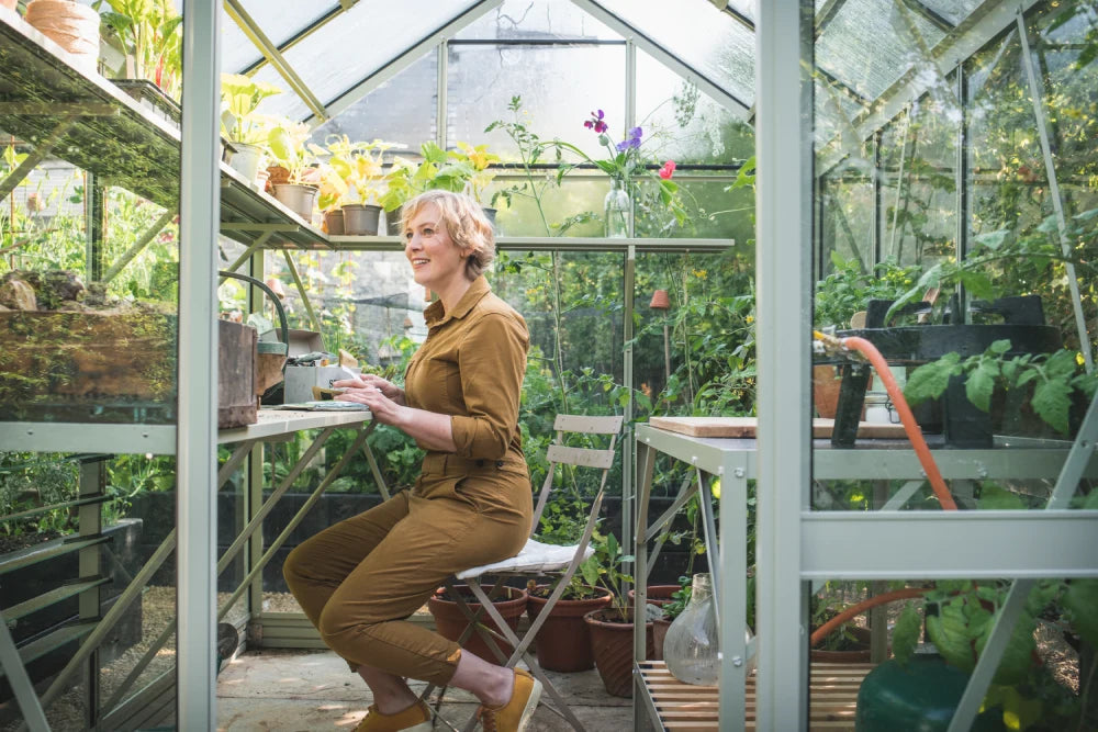 Woman sits at a table in a Rhino Greenhouse, arranging plants on shelves. Sunlight filters through the glass roof, surrounded by pots and gardening tools.