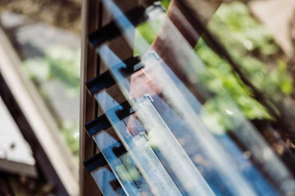 Glass panes form a louvered window, slightly open, in a Rhino Greenhouse setting. Sunlight filters through, illuminating the green foliage outside, creating a bright, airy atmosphere.
