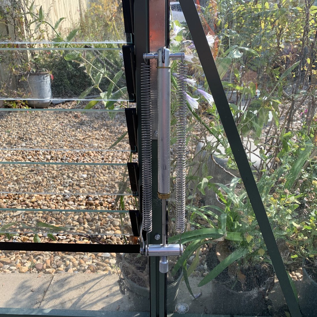 Metallic spring mechanism adjusting glass louver windows in a Rhino Greenhouse, surrounded by lush plants and gravel-filled ground, with sunlight filtering through the structure.
