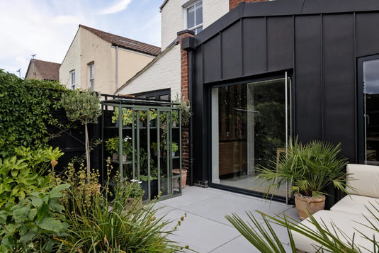 A small Rhino Greenhouse filled with plants stands next to a modern black extension of a house. Surrounded by lush greenery and potted plants, the setting is a stylish patio.