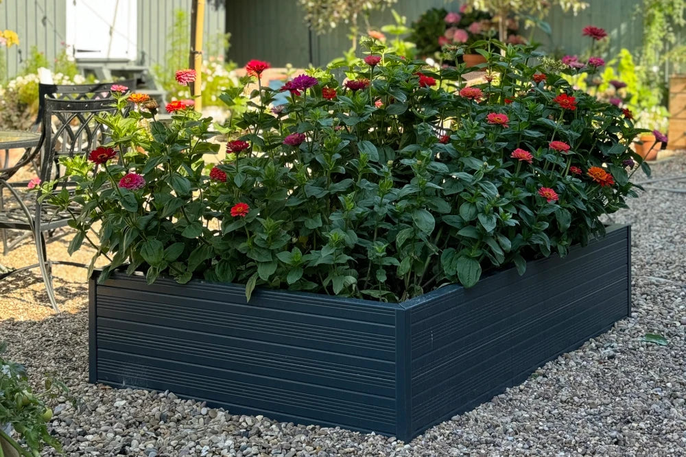 A dark gray raised planter box holds vibrant red and orange flowers, situated on gravel beside a metal garden chair and table set, with a garden backdrop.