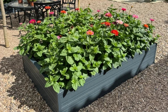A Rhino metal raised garden bed filled with vibrant flowering plants sits on a gravel surface. Nearby, a metal outdoor table and chairs suggest a garden seating area.