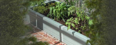A wooden planter box is filled with various green plants and soil, positioned on a brick patio, surrounded by blurred greenery.