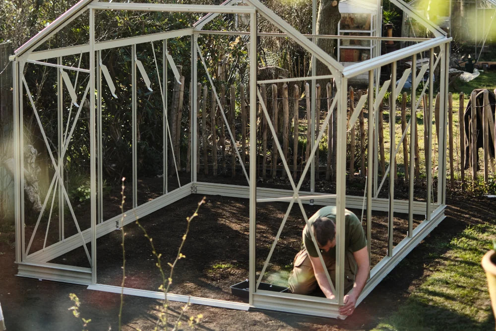 Rhino Greenhouse frame being assembled by a person kneeling, set in a garden with surrounding wooden fence and greenery.