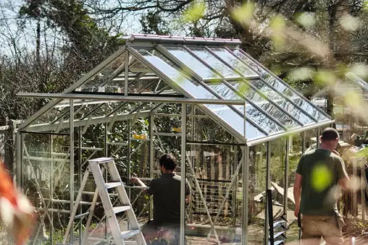 A metal-framed Rhino Greenhouse is being assembled; one person stands on a ladder, another nearby. Surrounded by trees and outdoor foliage, sunlight reflects off the glass panes.