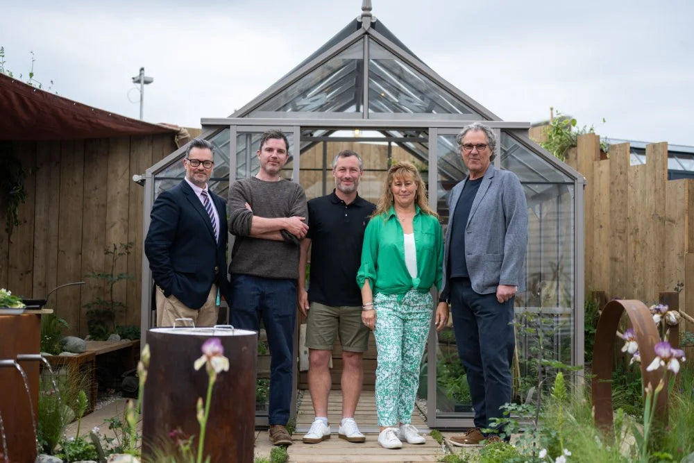Five people stand in front of a glass Rhino Greenhouse, posing for a photo. The setting is a garden area with wooden fences and decorative plants surrounding the structure.