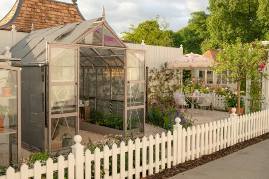 A small Rhino Greenhouse stands with open doors, revealing plants inside, surrounded by a white picket fence. Adjacent is a garden with a table, chairs, and potted flowers under an umbrella.