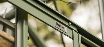 Rhino Greenhouse frame with Rhino label attached, situated outdoors among blurred greenery, suggesting a robust structure for gardening.